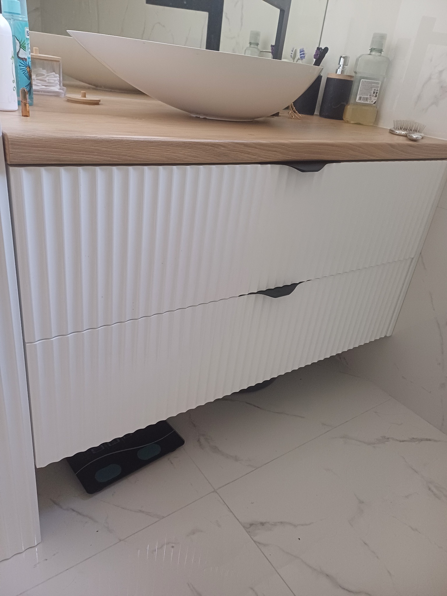 Nowoczesna szafka łazienkowa z białą, ryflowaną fasadą i drewnianym blatem, na którym stoi owalna umywalka i akcesoria. Na podłodze waga łazienkowa.