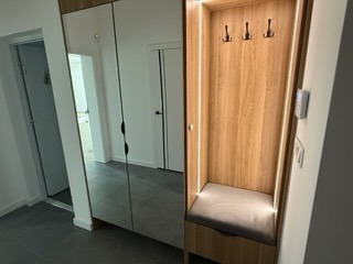 Nowoczesna szafa wnękowa z lustrzanymi drzwiami i oświetlonym wnęką siedziskiem z wieszakami. Jasne drewno, minimalistyczny design, widok na korytarz.