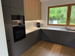 Nowoczesna kuchnia w odcieniach szarości z drewnianymi akcentami, oświetleniem LED i dużym oknem z widokiem na zieleń. Minimalistyczny design i funkcjonalne wyposażenie.