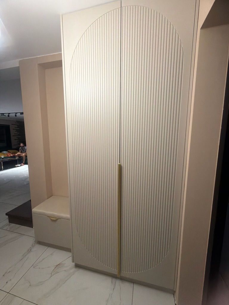 Beżowa szafa dwudrzwiowa z pionowymi żłobieniami i złotymi uchwytami, wkomponowana w przedpokój z siedziskiem. Minimalistyczny design, jasne wnętrze.