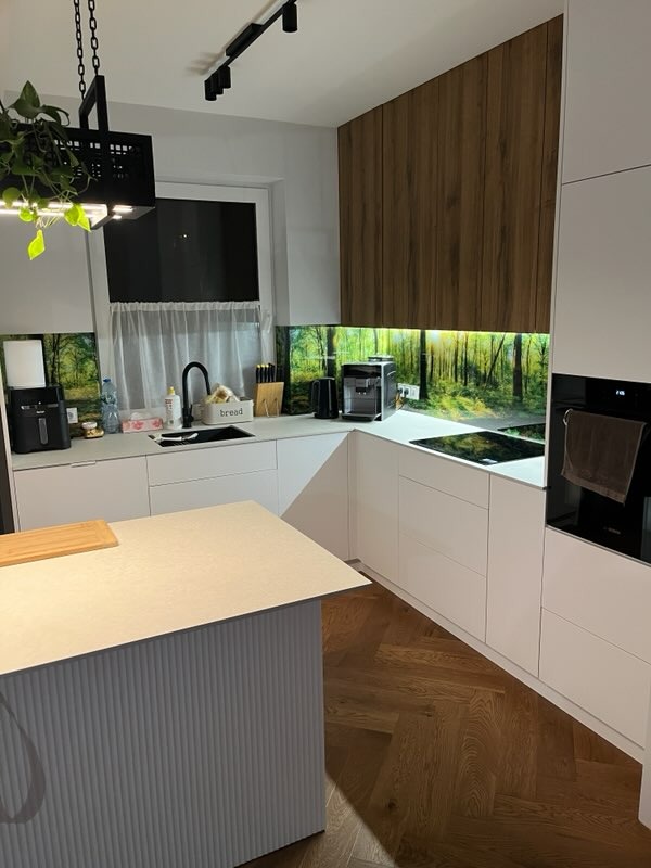 Biała kuchnia z wyspą i drewnianymi akcentami. Nad blatem panel z motywem leśnym. Parkiet w jodełkę. Nowoczesne oświetlenie punktowe.