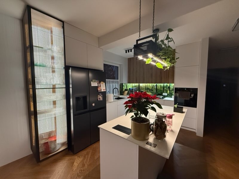 Nowoczesna kuchnia z wyspą, czarną lodówką i przeszkloną szafką z ryflowanego szkła. Nad wyspą designerska lampa z roślinami. Na blacie świąteczna gwiazda betlejemska.