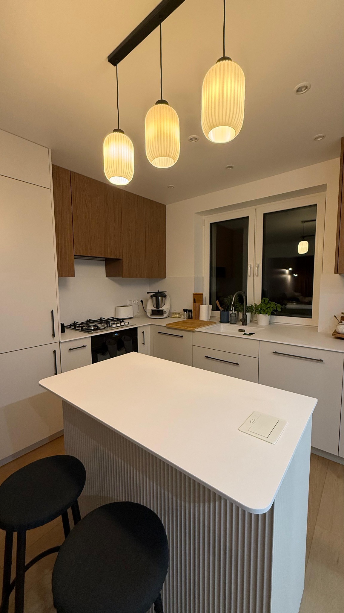 Nowoczesna kuchnia z wyspą i trzema lampami wiszącymi. Białe i drewniane szafki, czarne stołki barowe. Minimalistyczny design, ciepłe oświetlenie.