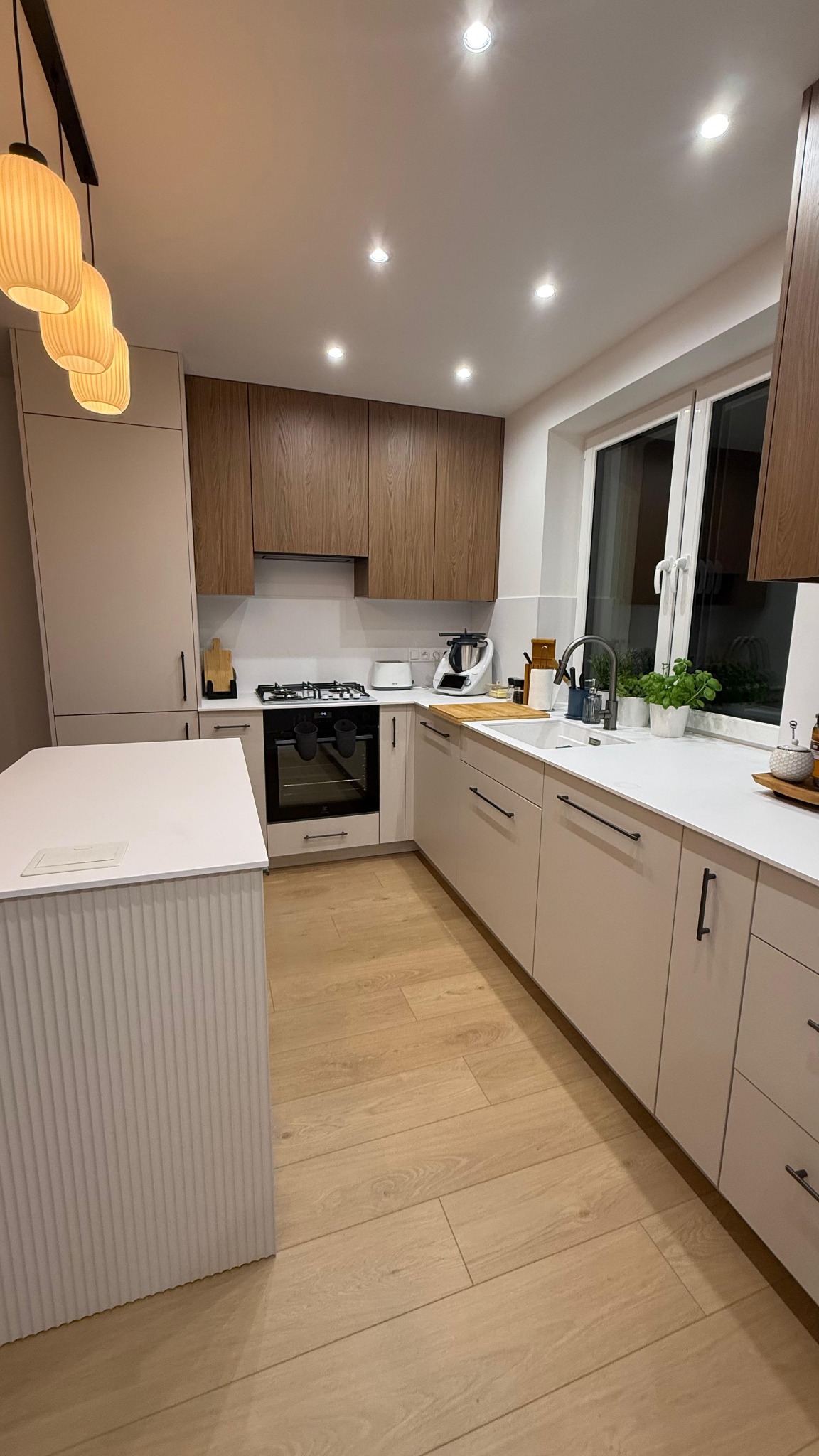 Jasna kuchnia z wyspą, fronty szafek w odcieniach beżu i drewna, czarne uchwyty, drewniana podłoga, oświetlenie punktowe i wiszące lampy.