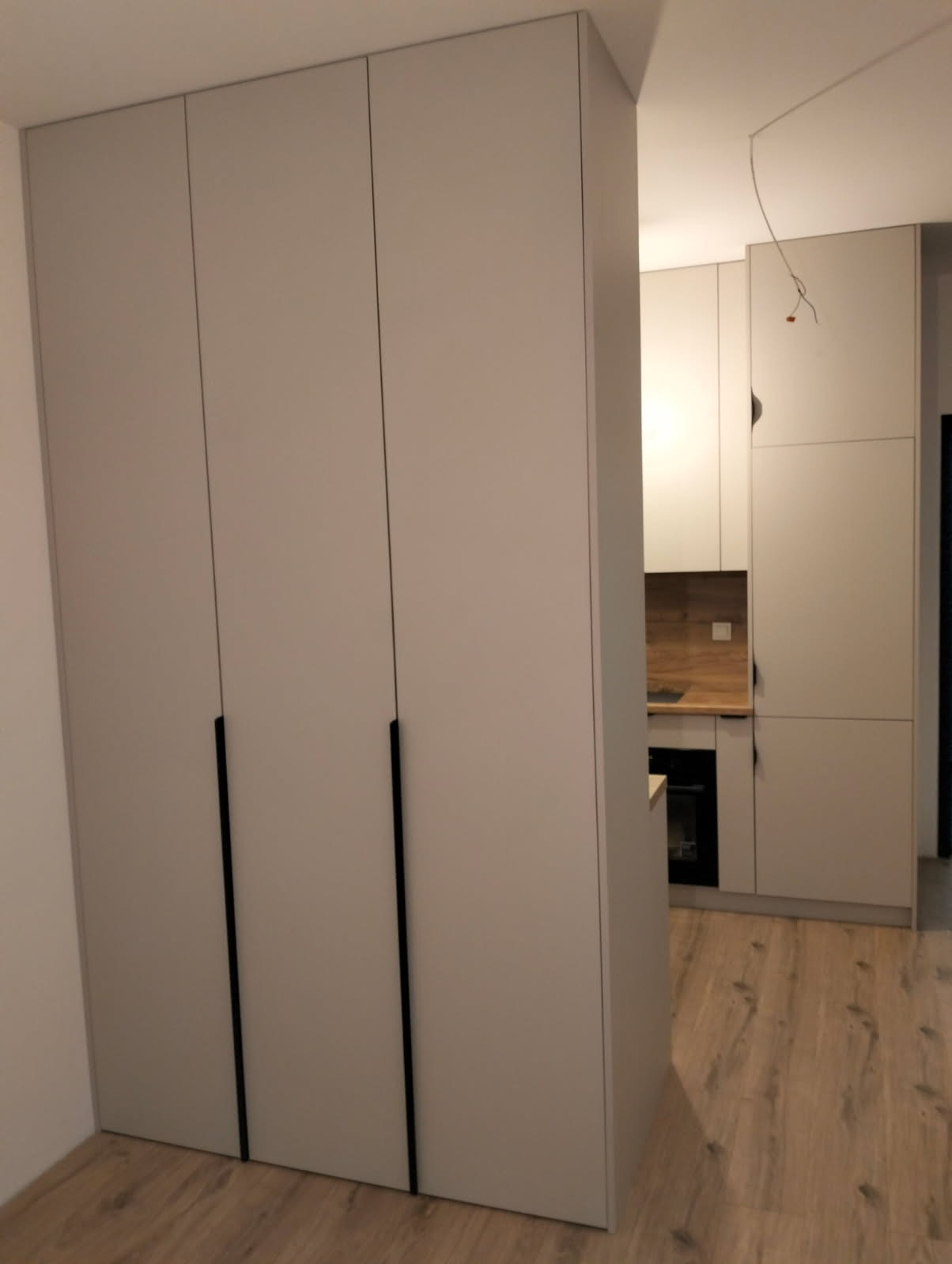 Jasnoszara szafa na wymiar z czarnymi uchwytami, widoczna z boku, z fragmentem zabudowy kuchennej w tle. Podłoga z paneli imitujących drewno.