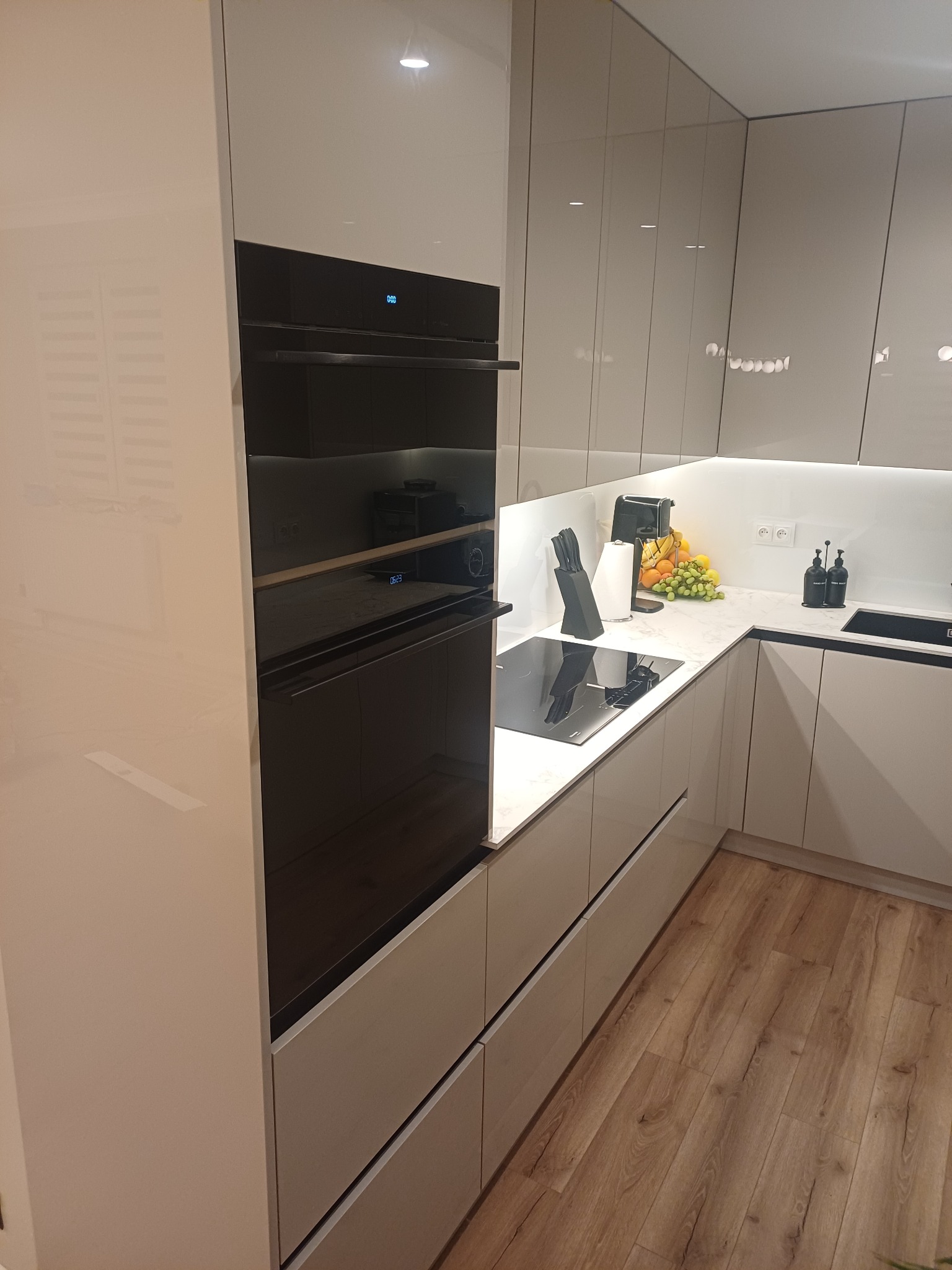 Nowoczesna kuchnia z zabudową w odcieniach szarości, z piekarnikiem w zabudowie i drewnianą podłogą. Minimalistyczny design z oświetleniem LED pod szafkami.