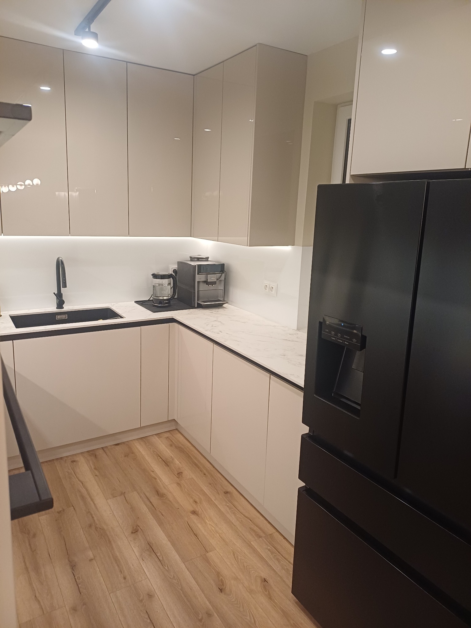 Jasna kuchnia z połyskiem: białe szafki, czarny zlew i armatura, marmurowy blat, ekspres do kawy i czarna lodówka side-by-side. Drewniana podłoga.