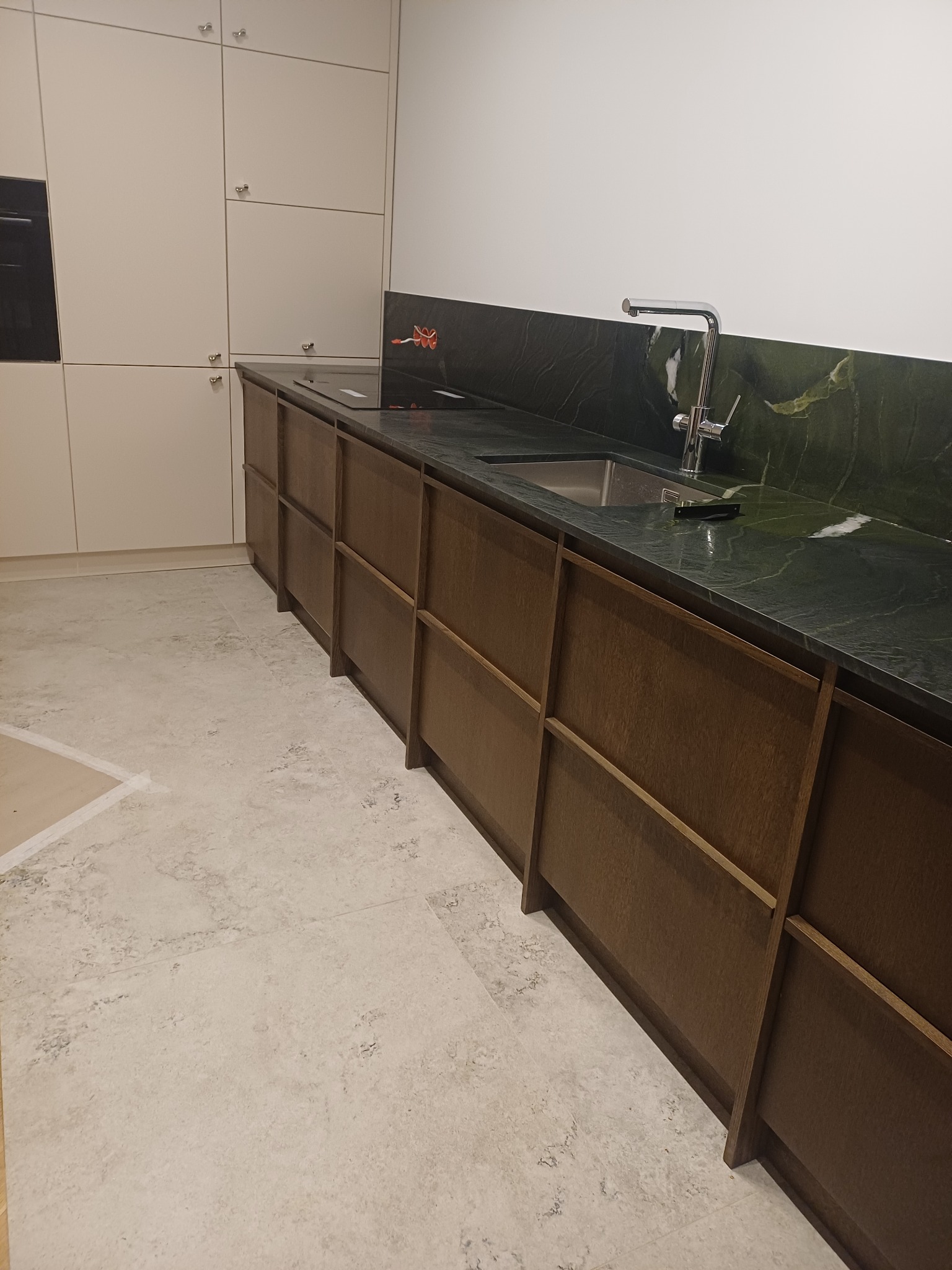 Nowoczesna kuchnia z drewnianymi szafkami i blatem z zielonego marmuru. Minimalistyczny design, chromowana bateria. Widoczne fragmenty podłogi z jasnych płytek.