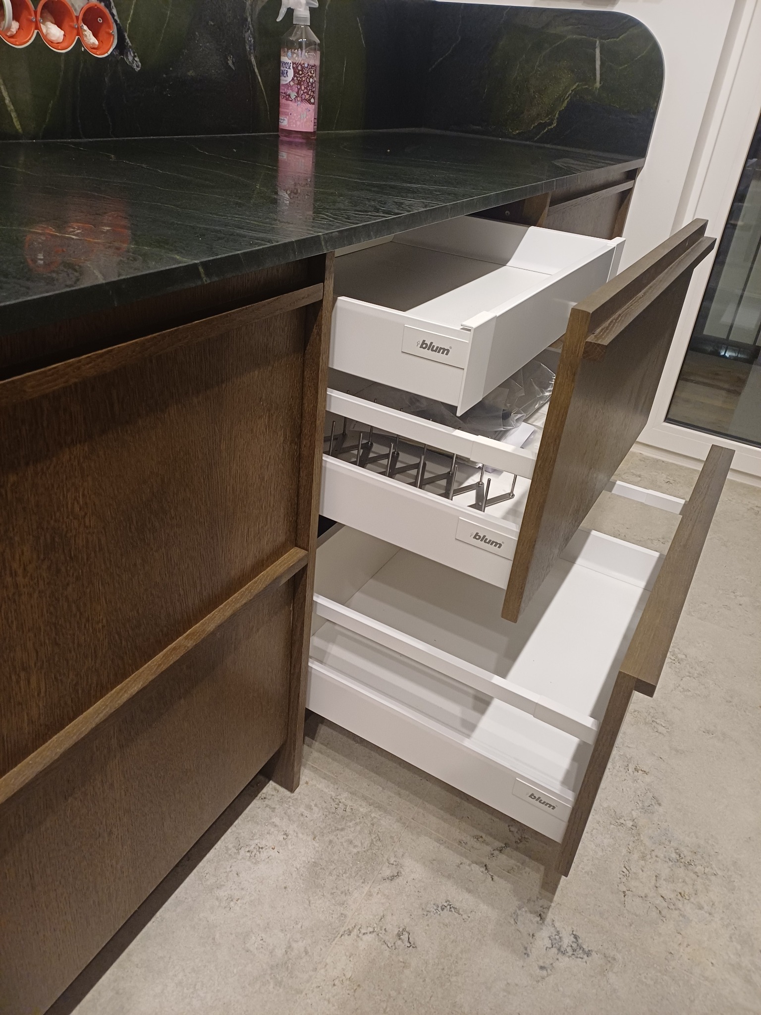 Otwarta szafka kuchenna z szufladami Blum, widoczne wewnętrzne organizery i ciemny blat z zielonymi żyłkami. Fronty szafek w odcieniach brązu.