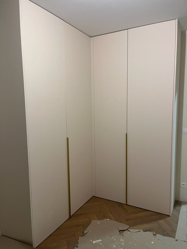 Jasna, narożna szafa wnękowa z długimi, złotymi uchwytami, na parkiecie ułożonym w jodełkę. Widoczne ślady po remoncie.