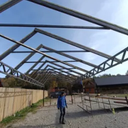 Metalowa konstrukcja dachu w trakcie budowy, widok z dołu na niebo. Na pierwszym planie osoba w niebieskiej kurtce, w tle drewniany budynek.