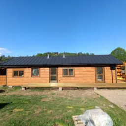 Gotowy, parterowy dom z bali z ciemnym, metalowym dachem i drewnianym tarasem, otoczony zielenią. Widok na elewację frontową w słoneczny dzień.