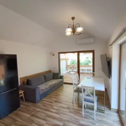 Jasne wnętrze z drewnopodobną podłogą: sofa, stół z krzesłami, czarna lodówka, wyjście na balkon z białymi meblami ogrodowymi. Widok z perspektywy.