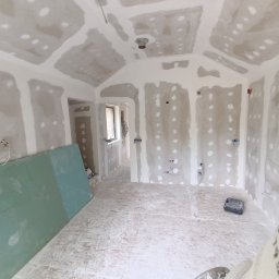 Dom-bud Serhii Varetskji - Wnętrze w trakcie wykańczania: sufit i ściany pokryte płytami gipsowo-kartonowymi, widoczne łączenia szpachlowane. Na podłodze płyty OSB, w tle okno i instalacje.
