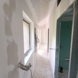 Dom-bud Serhii Varetskji - Korytarz w trakcie wykańczania: ściany z płyt karton-gips, widoczne przewody elektryczne, surowy stan. Okno z zasłoniętą szybą. Metalowe futryny drzwi.