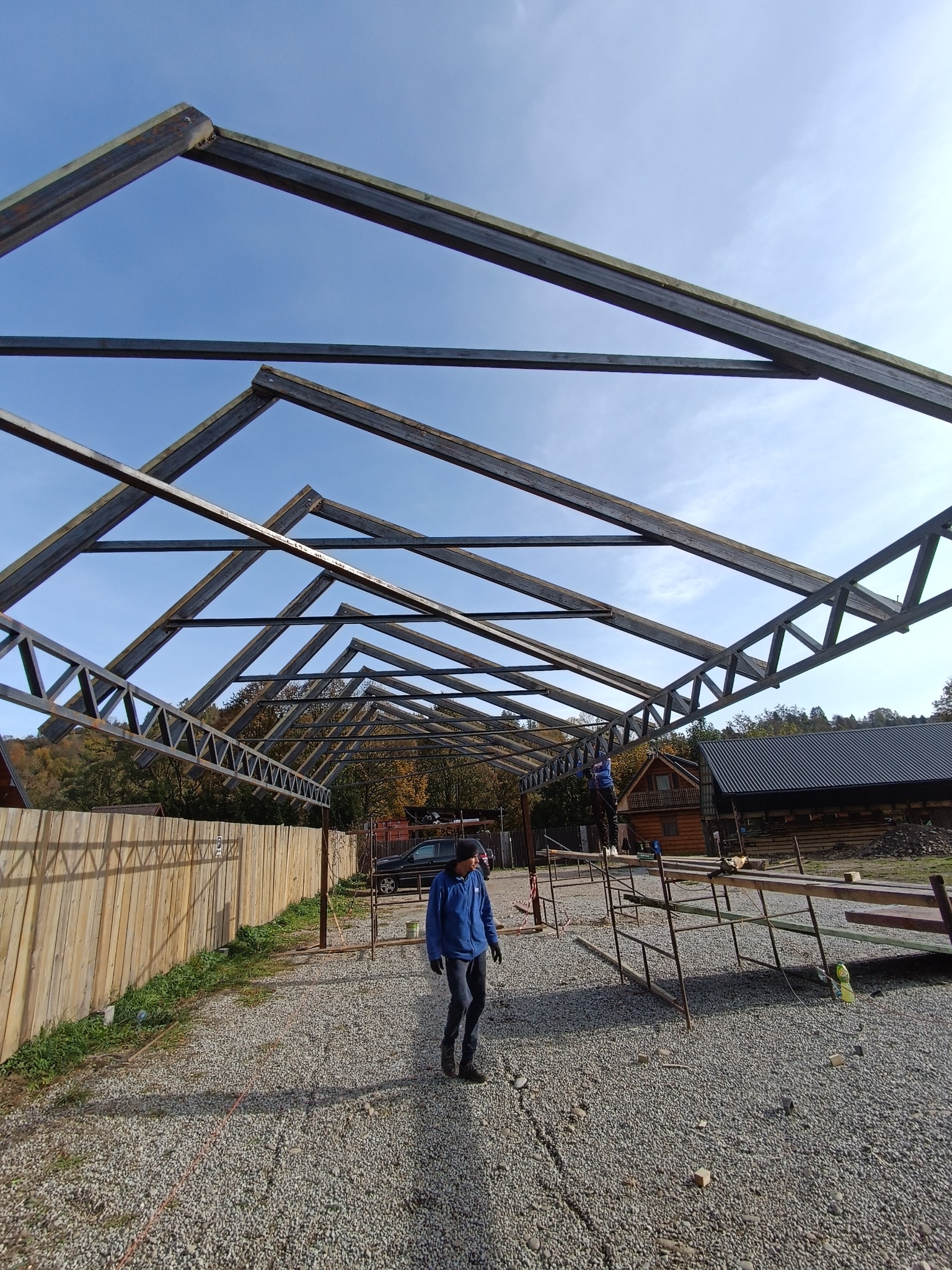 Metalowa konstrukcja dachu w trakcie budowy, widok z dołu na niebo. Na pierwszym planie osoba w niebieskiej kurtce, w tle drewniany budynek.