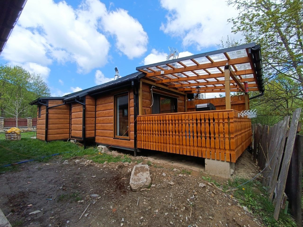 Drewniany dom z tarasem i przezroczystym zadaszeniem w słoneczny dzień. Widoczne detale wykończenia i ogrodzenie. Ujęcie z niskiej perspektywy.