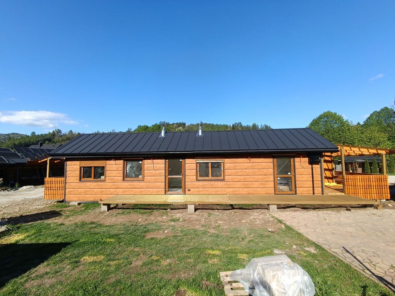 Gotowy, parterowy dom z bali z ciemnym, metalowym dachem i drewnianym tarasem, otoczony zielenią. Widok na elewację frontową w słoneczny dzień.