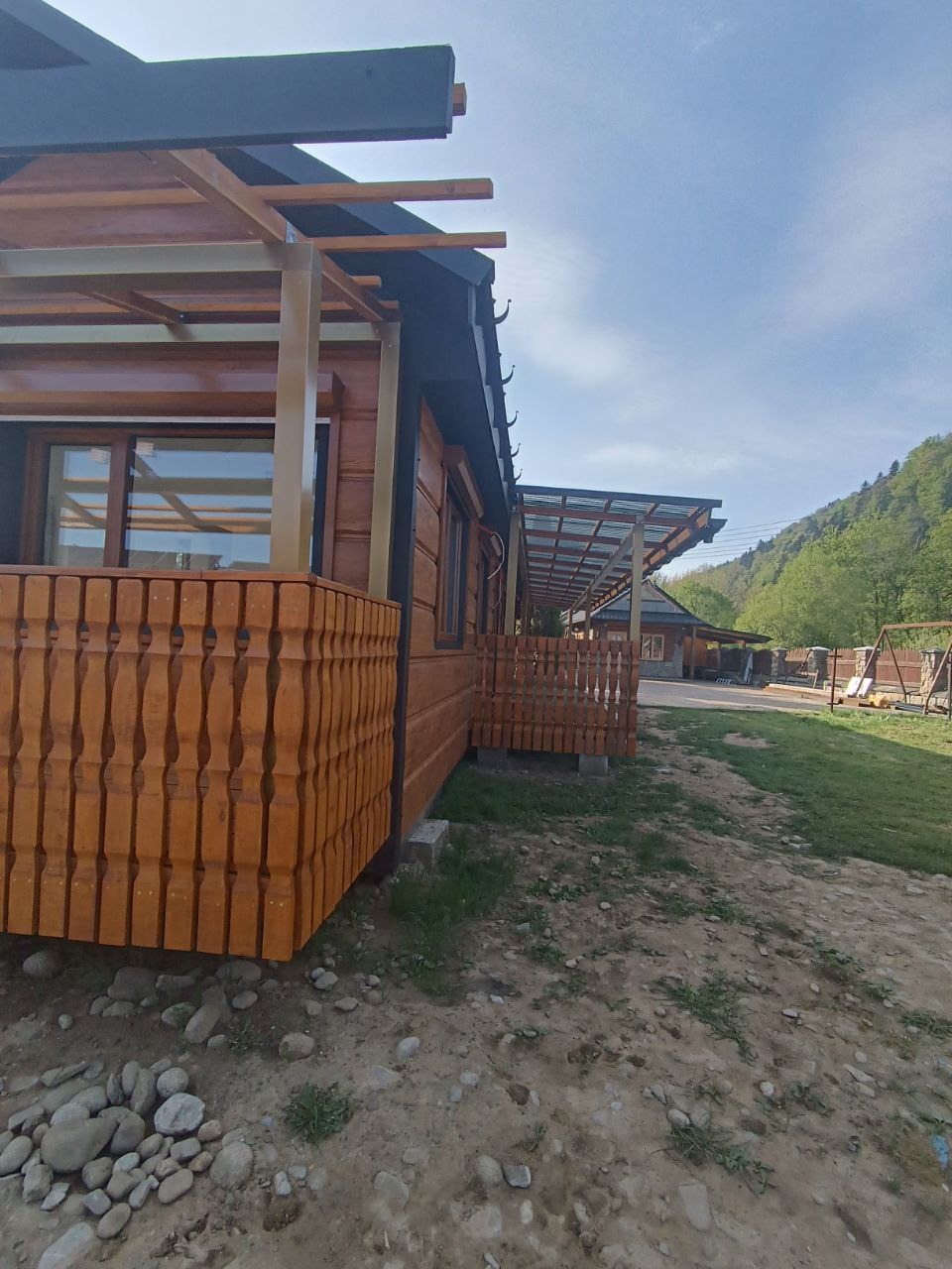 Drewniany dom z osłoniętym tarasem i czarnym dachem, na tle zielonych wzgórz. Widok z dołu, podkreślający konstrukcję dachu i wykończenie elewacji.