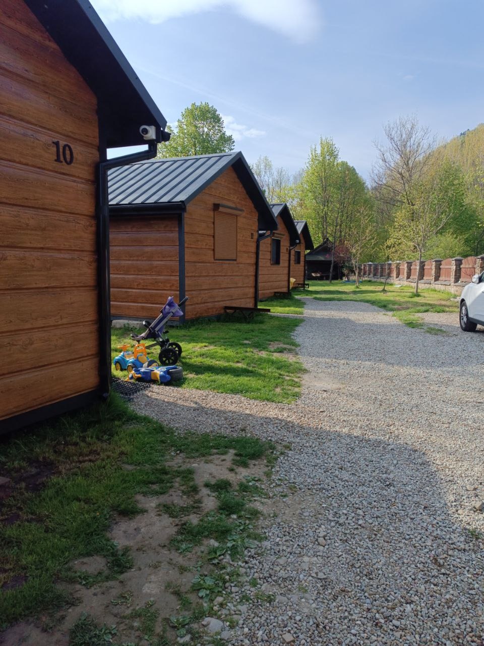 Rząd drewnianych domków letniskowych z numerem 10 na pierwszym planie, otoczonych zielenią i żwirową ścieżką, z widocznym w tle zalesionym wzgórzem.