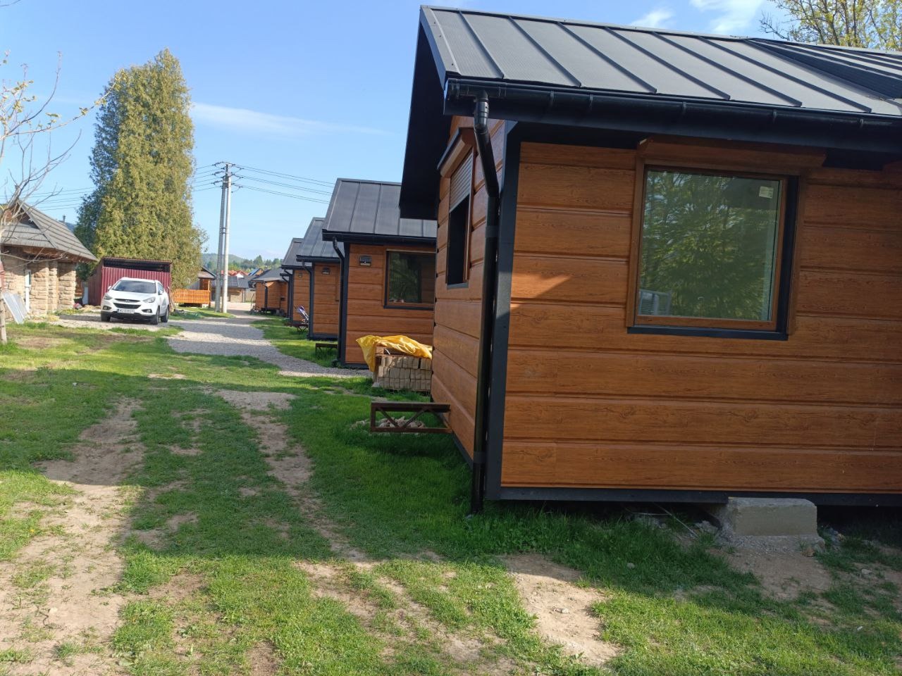 Szereg brązowych domków letniskowych z szarym dachem, widocznych z perspektywy trawnika. W tle drzewa i biały samochód. Słoneczny dzień.