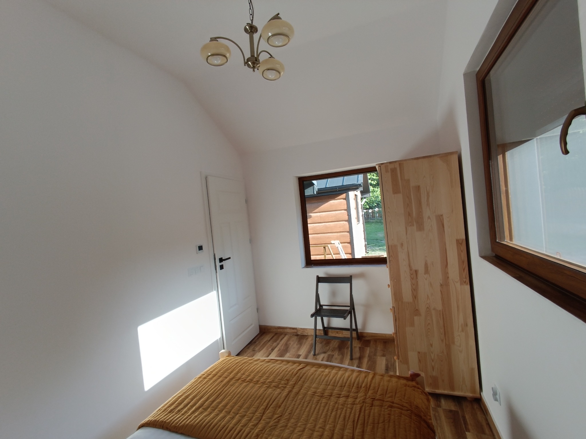 Jasne wnętrze pokoju z drewnianą podłogą i szafą w Krościenku nad Dunajcem. Widoczne okno, drzwi i łóżko z pomarańczową narzutą. Uwagę zwraca lampa sufitowa.