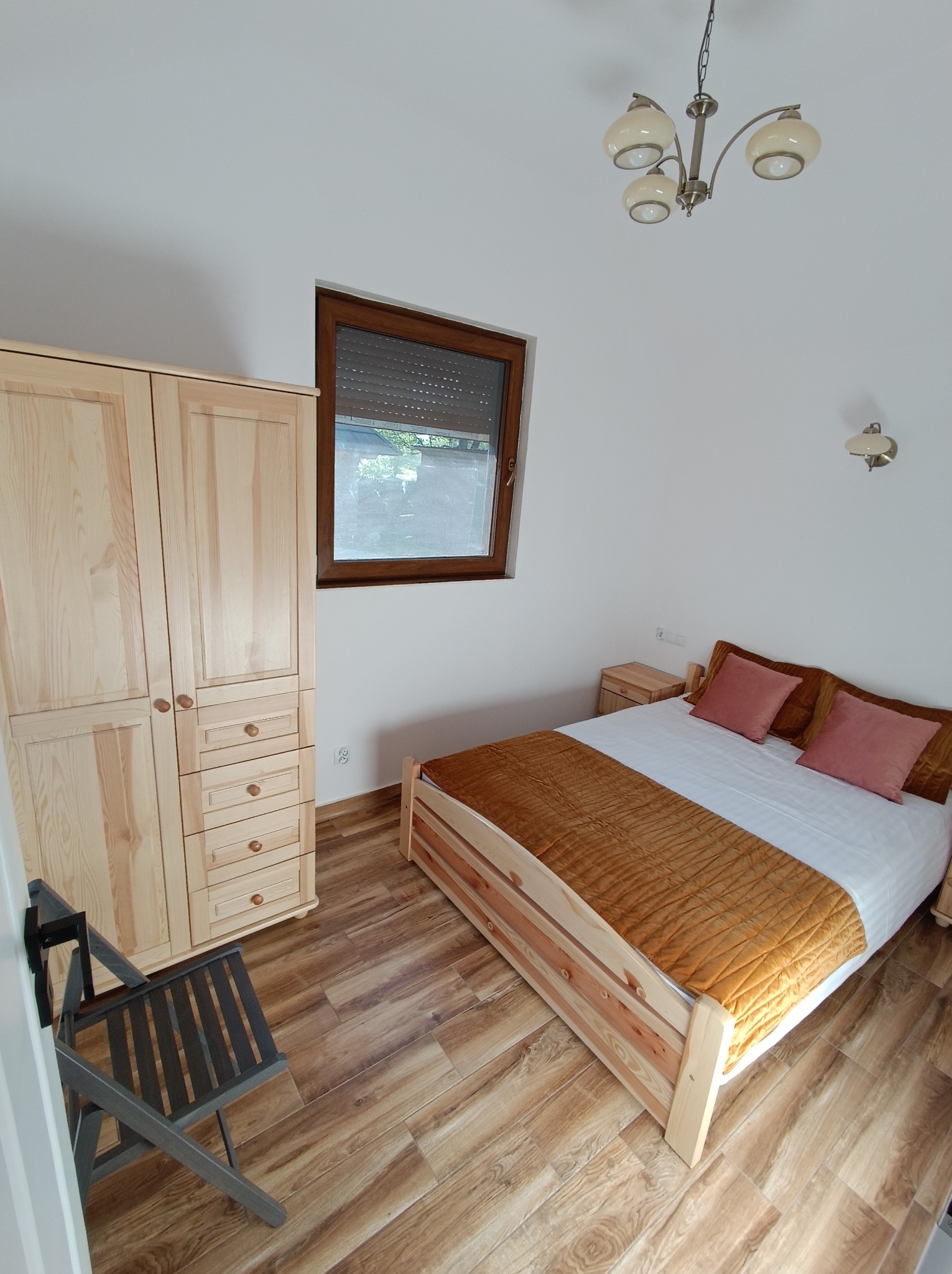 Jasne drewniane panele podłogowe w sypialni z łóżkiem, szafą i krzesłem. Widok z góry na wnętrze z oknem i lampą sufitową.