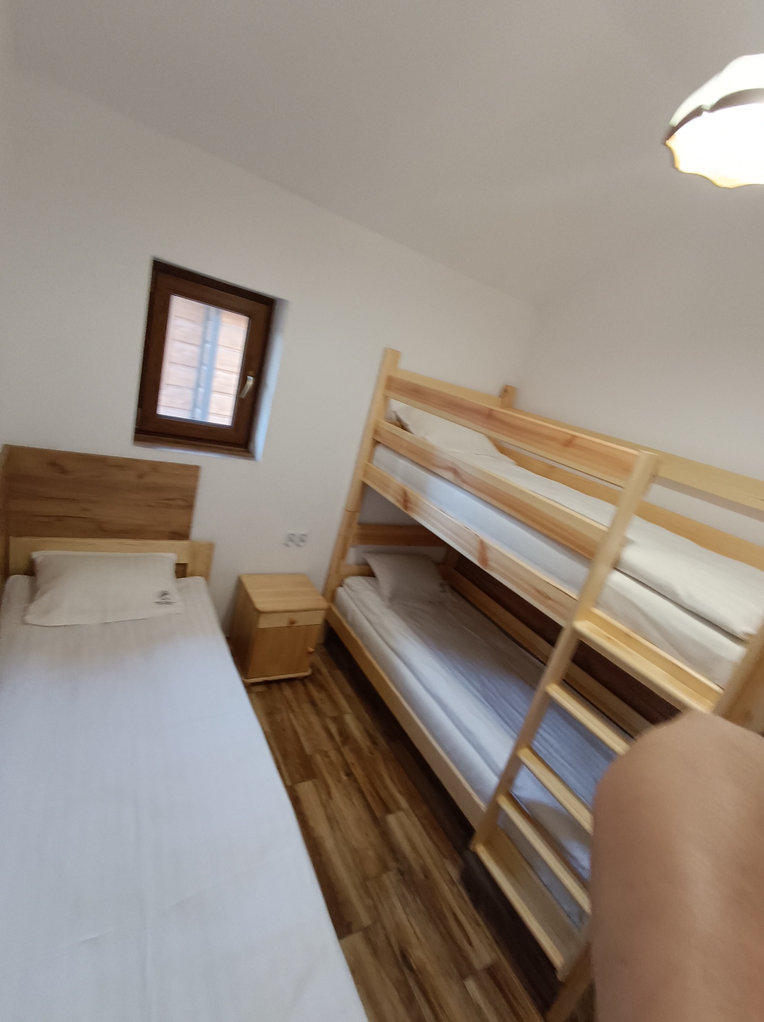 Jasne wnętrze pokoju z drewnianymi łóżkami piętrowymi i pojedynczym łóżkiem. Widoczne okno i drewniana podłoga. Minimalistyczny wystrój.