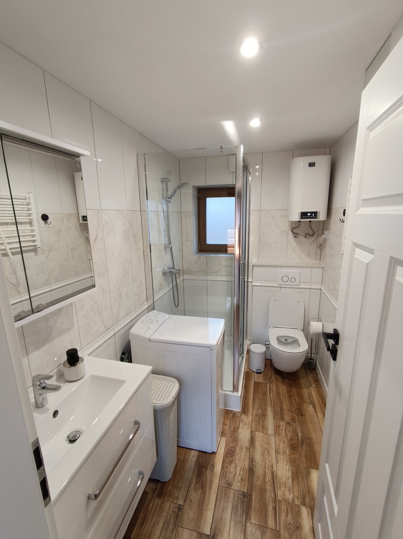 Jasna łazienka z drewnopodobną podłogą, kabiną prysznicową, pralką i umywalką z szafką. Białe płytki z delikatnym wzorem marmuru na ścianach.