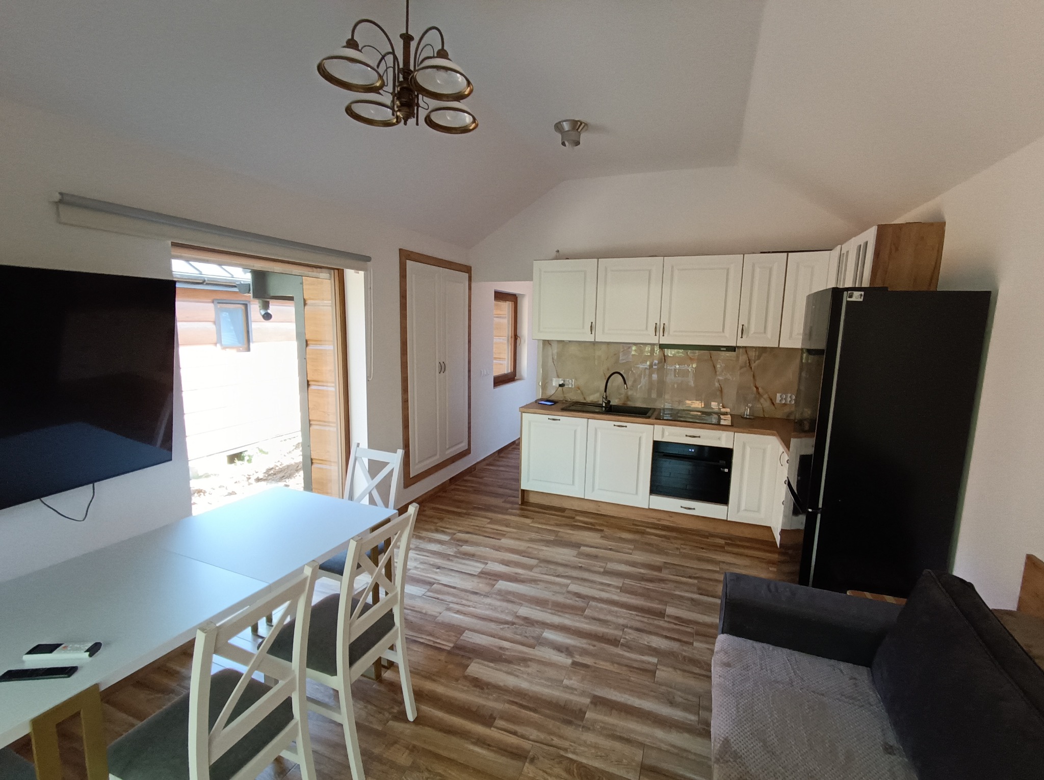 Jasne wnętrze domu z drewnianą podłogą, białymi meblami kuchennymi i stołem. Widoczny fragment sofy i okno z widokiem na zewnątrz. Całość doświetlona przez lampę sufitową.