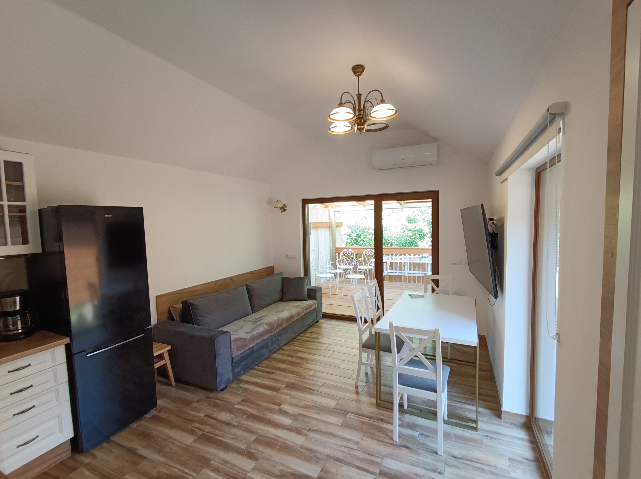 Jasne wnętrze z drewnopodobną podłogą: sofa, stół z krzesłami, czarna lodówka, wyjście na balkon z białymi meblami ogrodowymi. Widok z perspektywy.