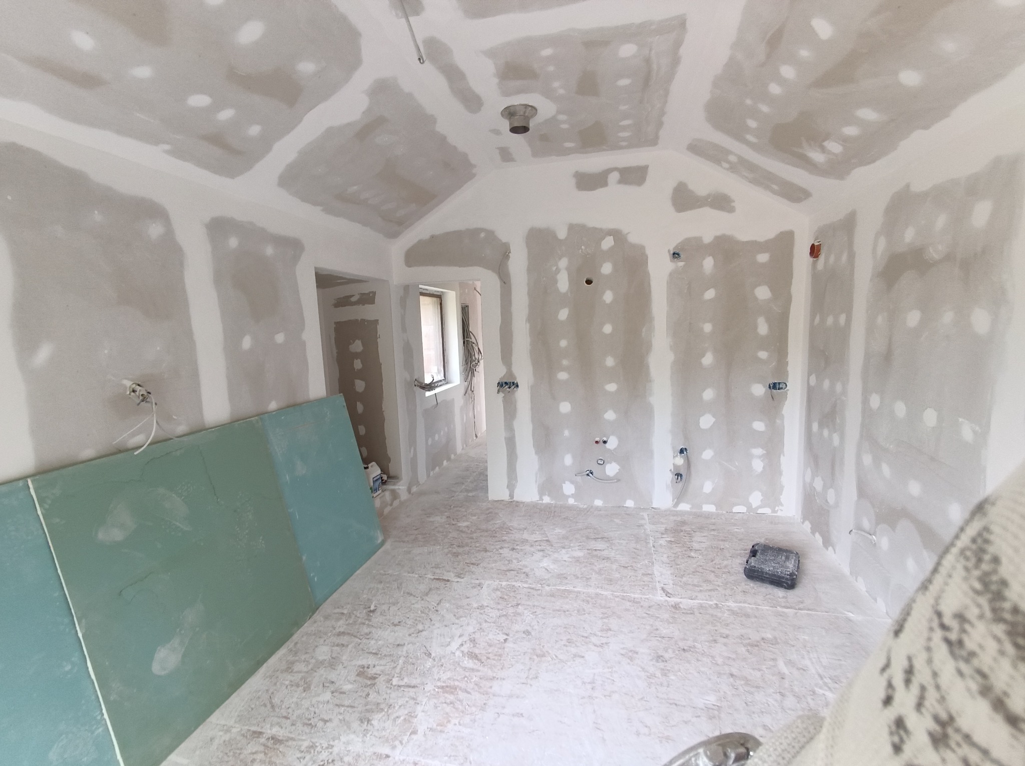 Wnętrze w trakcie wykańczania: sufit i ściany pokryte płytami gipsowo-kartonowymi, widoczne łączenia szpachlowane. Na podłodze płyty OSB, w tle okno i instalacje.