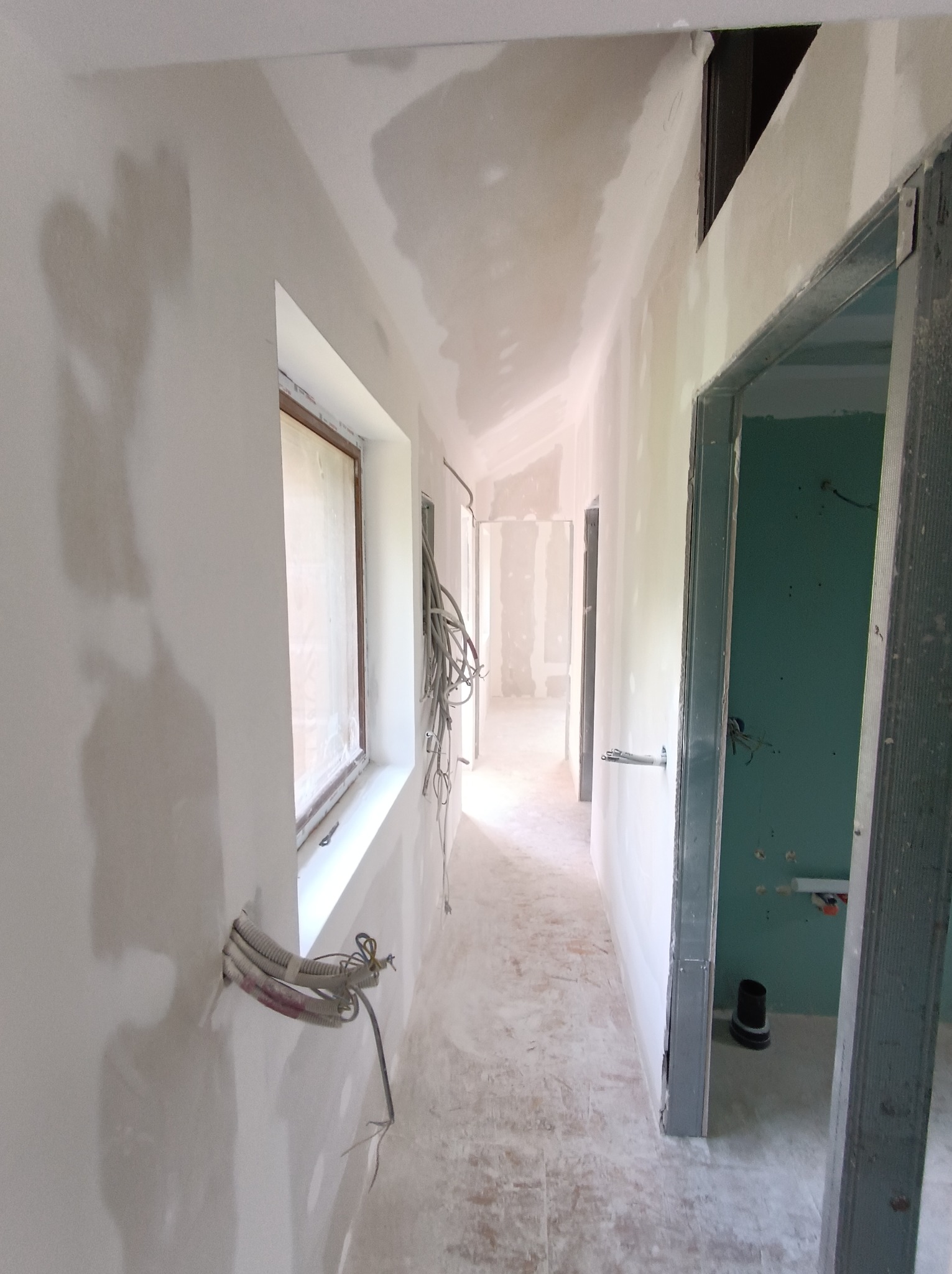 Korytarz w trakcie wykańczania: ściany z płyt karton-gips, widoczne przewody elektryczne, surowy stan. Okno z zasłoniętą szybą. Metalowe futryny drzwi.