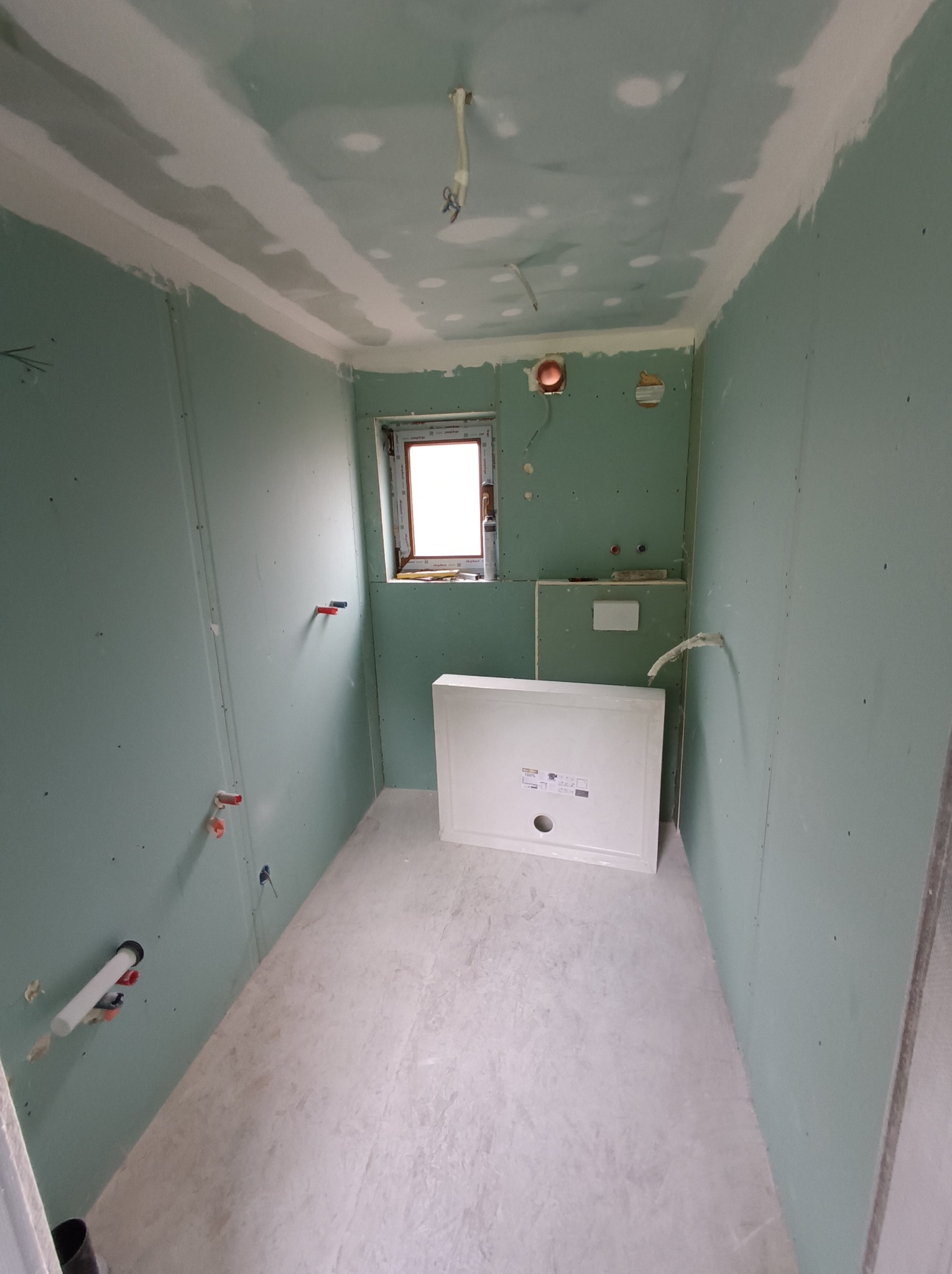 Wnętrze łazienki w trakcie wykańczania: ściany z płyt gipsowo-kartonowych, widoczne instalacje wodne, przygotowane miejsce na brodzik. Surowe, jasne światło.