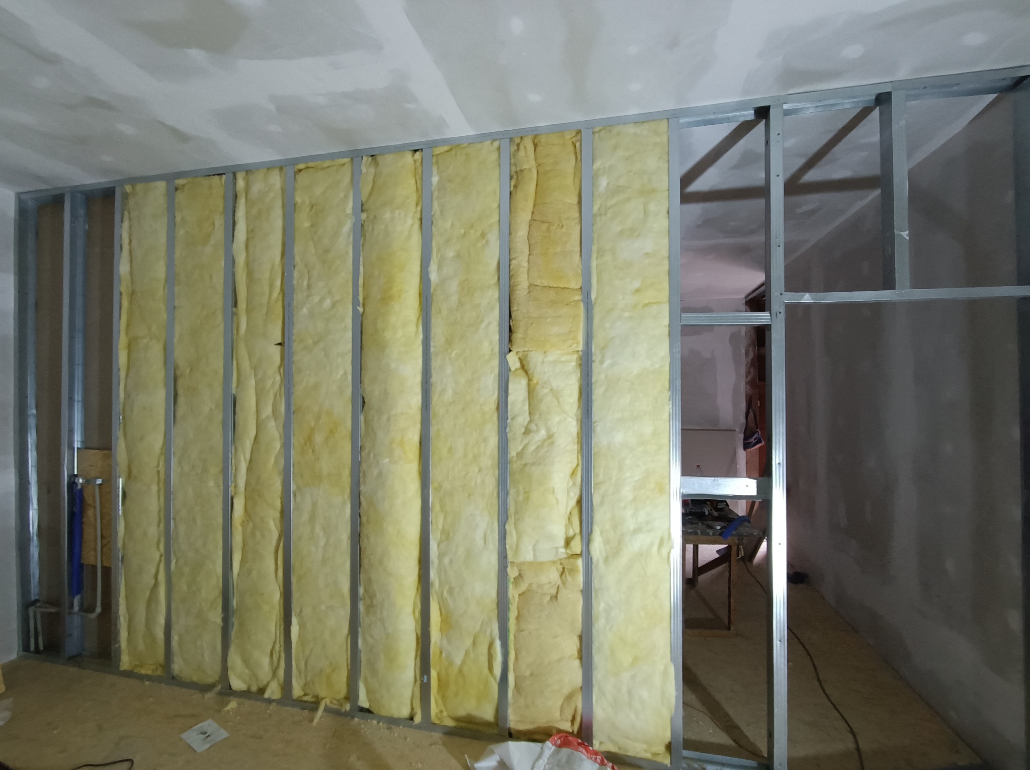 Konstrukcja ścianki działowej z metalowych profili wypełniona wełną mineralną. Widoczne instalacje, fragment podłogi i sufitu. Prace wykończeniowe w surowym wnętrzu.