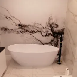 Nowoczesna łazienka z wolnostojącą białą wanną, czarną armaturą i marmurową tapetą na ścianie. Na sedesie leżą narzędzia, obok rolka papieru i pudełko z elementami montażowymi.