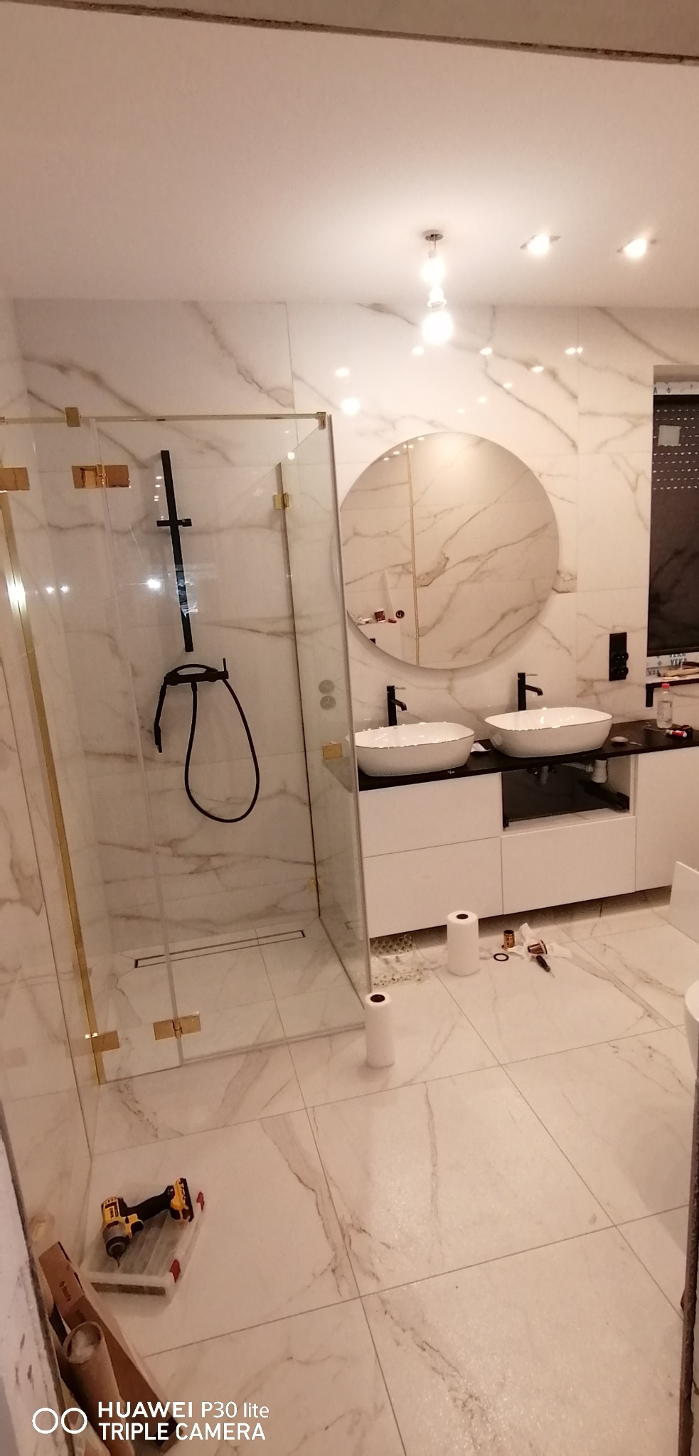 Nowoczesna łazienka z marmurowymi płytkami, kabiną prysznicową walk-in z czarną armaturą i złotymi detalami, okrągłym lustrem nad podwójną umywalką na białej szafce. Na podłodze narzędzia i rolki...