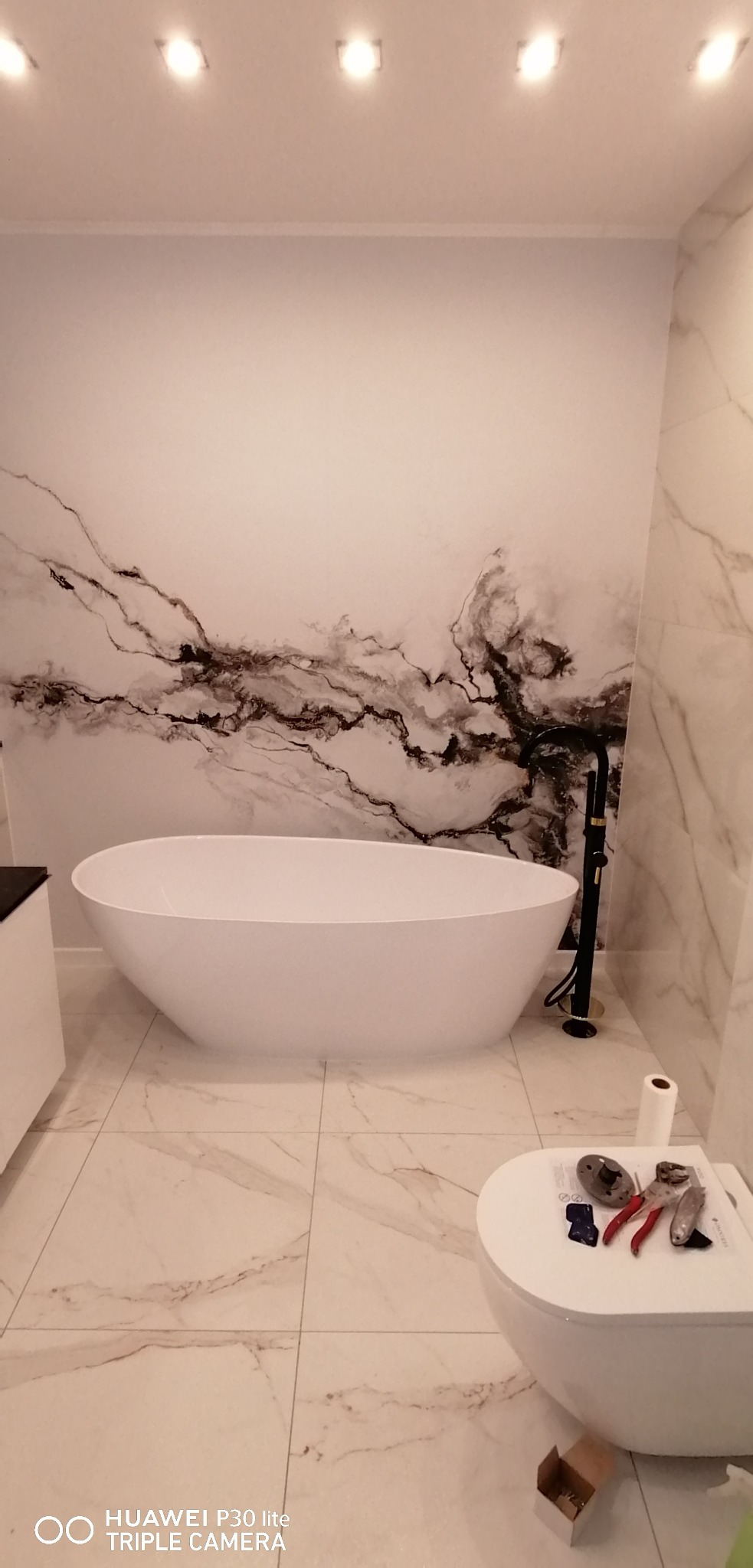 Nowoczesna łazienka z wolnostojącą białą wanną, czarną armaturą i marmurową tapetą na ścianie. Na sedesie leżą narzędzia, obok rolka papieru i pudełko z elementami montażowymi.