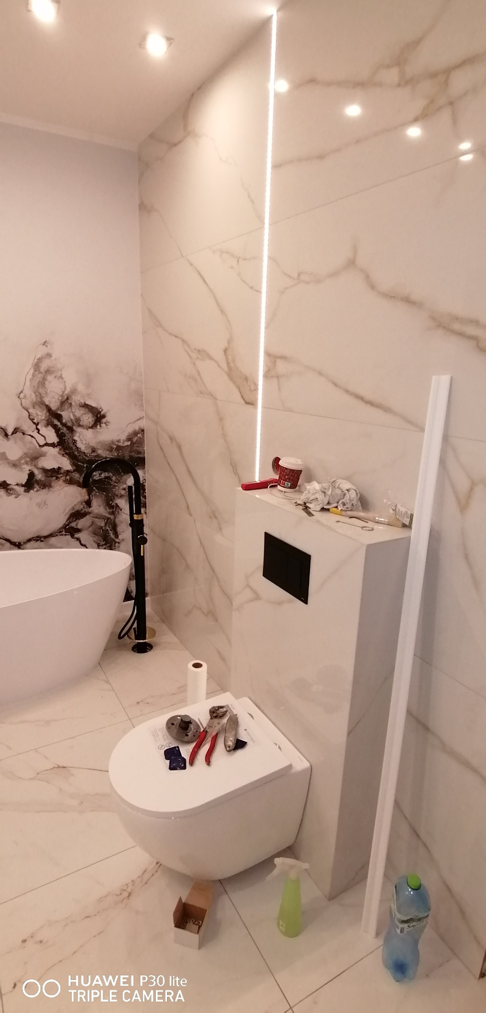 Nowoczesna łazienka w trakcie montażu: wisząca toaleta z narzędziami na desce, białe płytki imitujące marmur, czarna armatura i oświetlenie LED wzdłuż ściany.