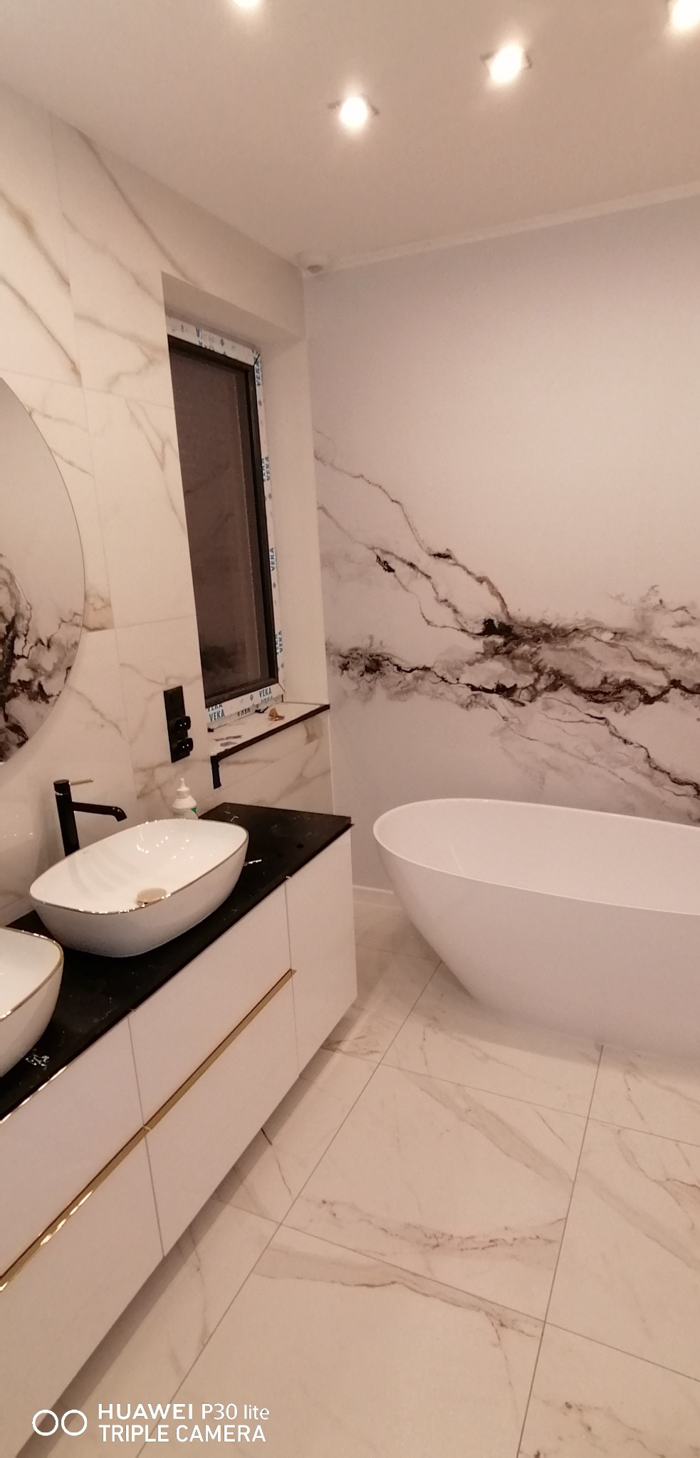 Nowoczesna łazienka z wolnostojącą białą wanną, umywalkami nablatowymi w kolorze białym ze złotymi akcentami, czarnym blatem, marmurowymi płytkami na ścianach i podłodze oraz oknem z ramą VEKA.