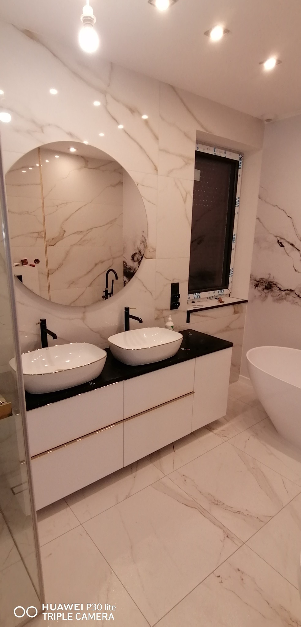 Nowoczesna łazienka z marmurowymi płytkami, okrągłym lustrem nad podwójną umywalką nablatową z czarną armaturą i białymi szafkami z złotymi akcentami.