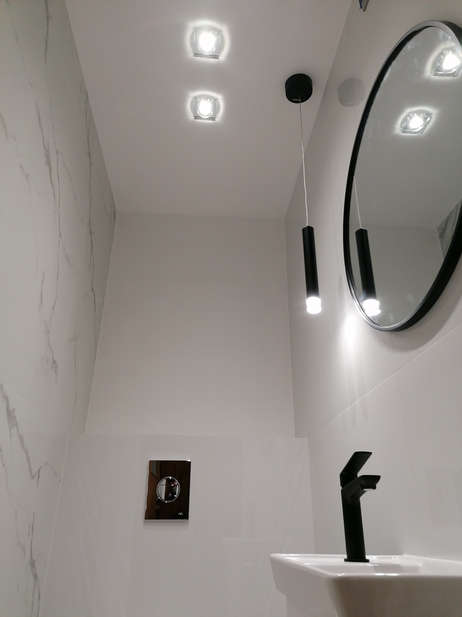 Nowoczesna łazienka w bieli i czerni: marmurowe płytki, okrągłe lustro, czarna bateria umywalkowa i wisząca lampa tubowa.