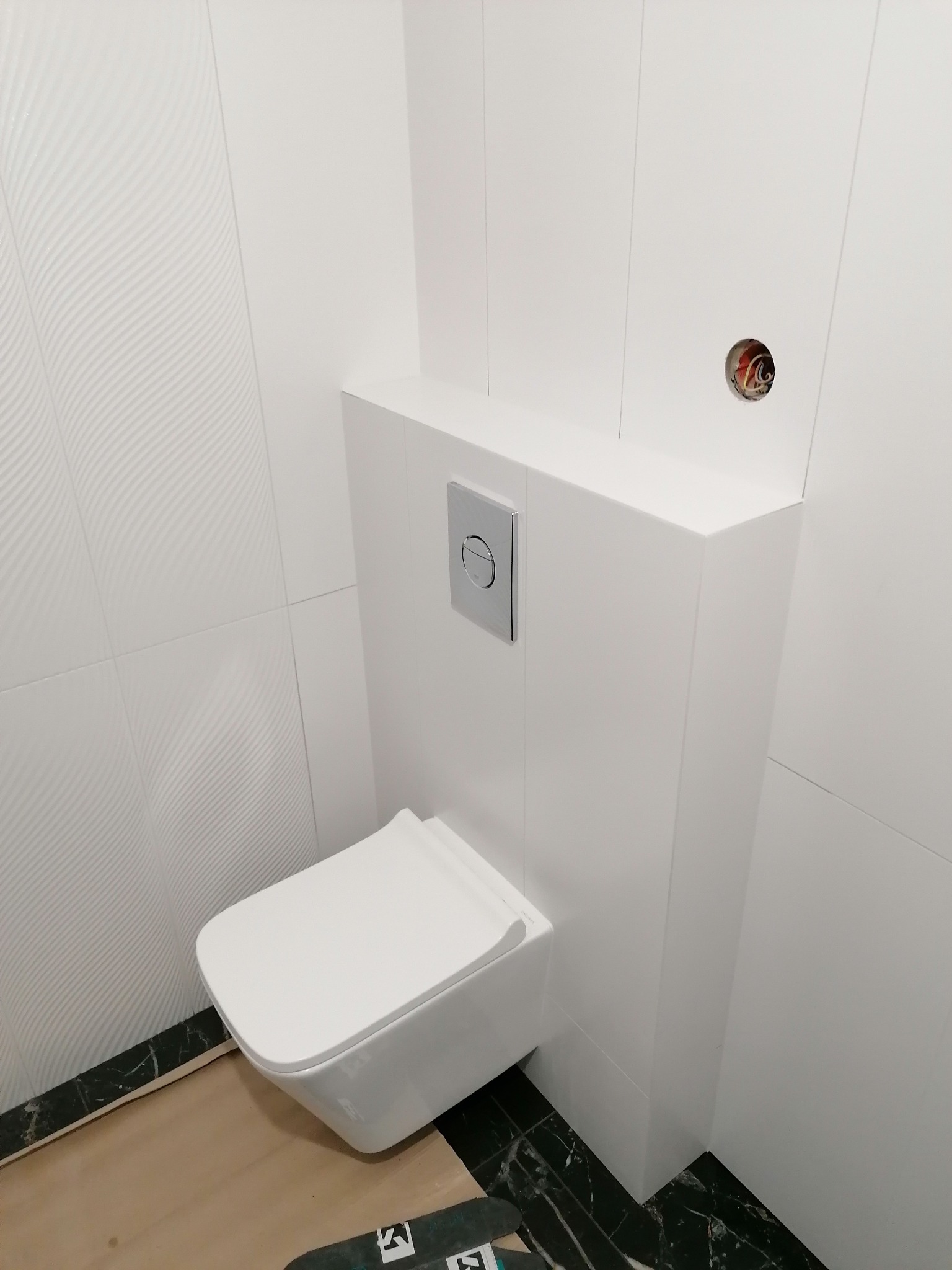 Nowoczesna, podwieszana toaleta z chromowanym przyciskiem spłukującym, osadzona na tle białych, wzorzystych płytek ściennych i marmurowej podłogi w trakcie prac wykończeniowych, widoczne...