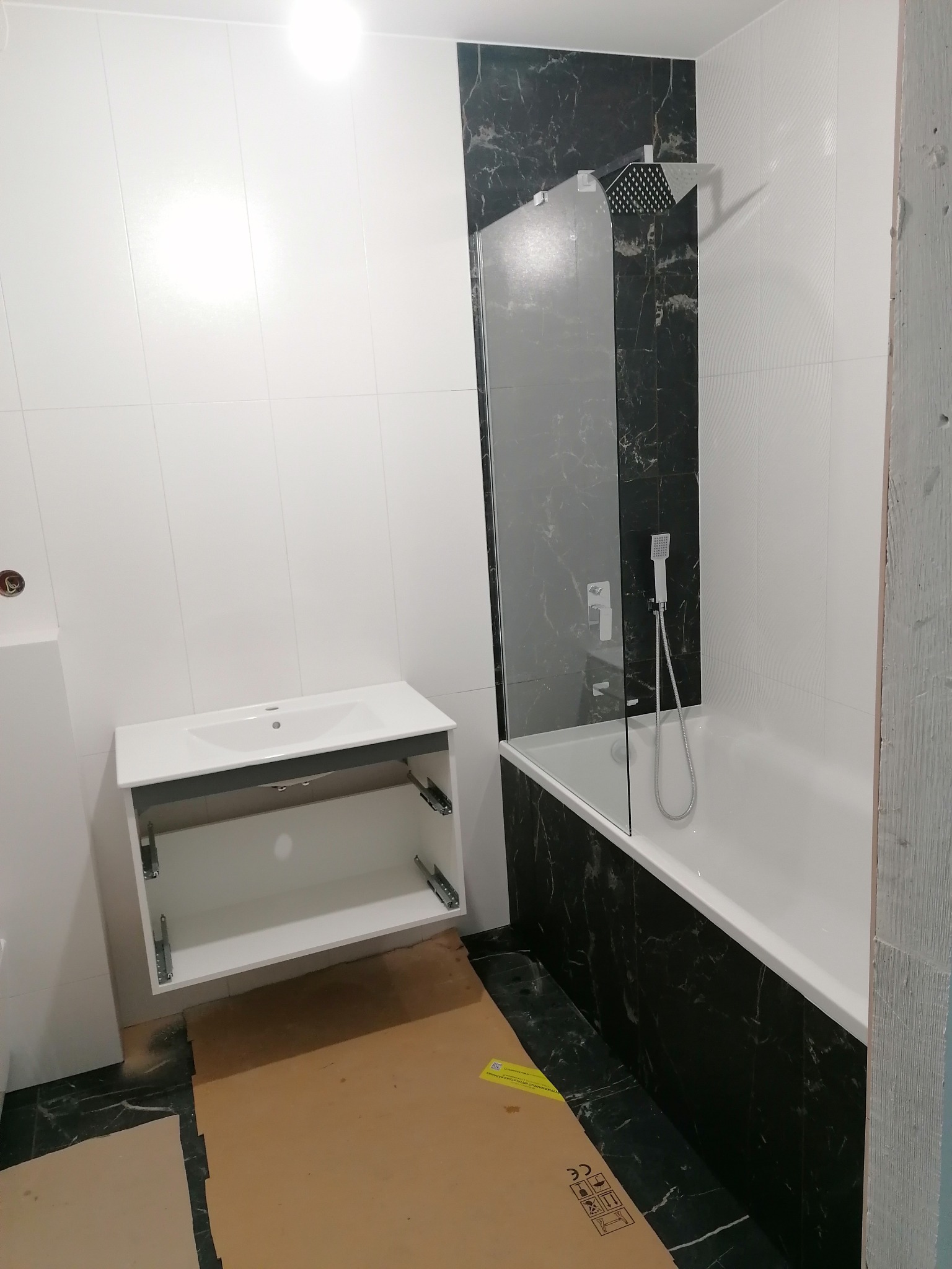 Wnętrze łazienki w trakcie montażu: białe płytki ścienne kontrastują z czarnymi płytkami imitującymi marmur wokół wanny z przeszklonym parawanem. Otwarta szafka pod umywalkę z widocznymi...