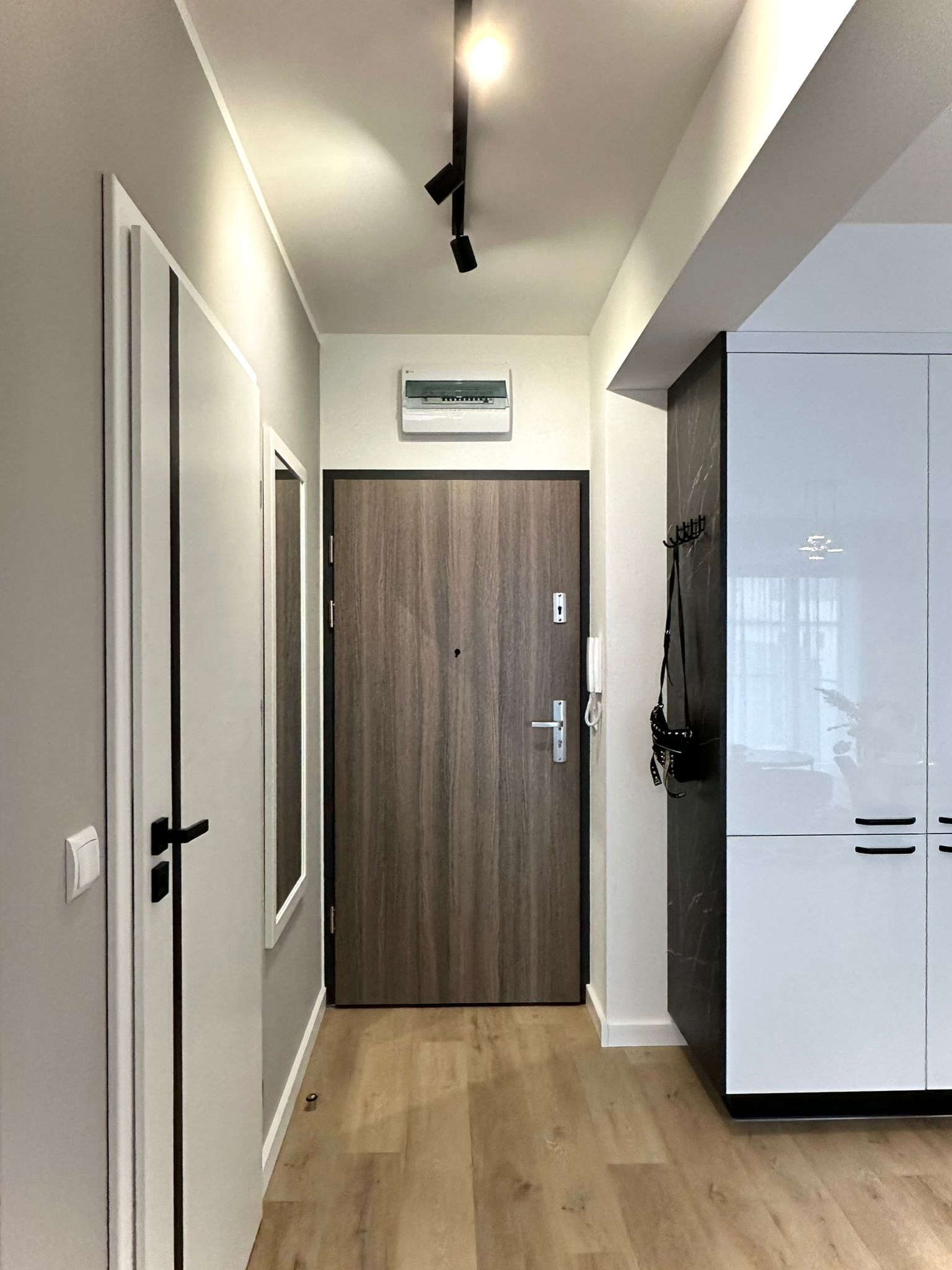 Nowoczesny przedpokój z drewnopodobną podłogą, szarą ścianą, białą szafą w zabudowie i drzwiami wejściowymi z wizjerem, oświetlony reflektorami.