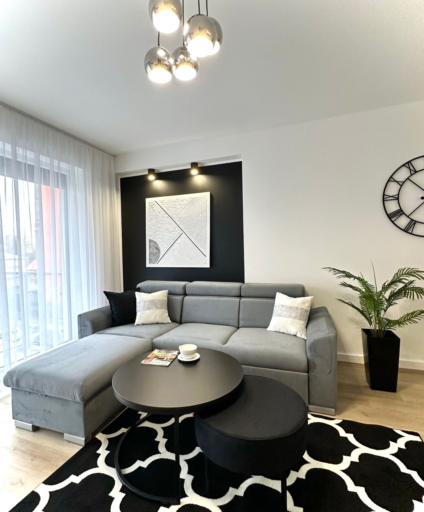Jasne, nowoczesne wnętrze salonu z szarą sofą narożną, czarnym stolikiem kawowym, wzorzystym dywanem, rośliną doniczkową i dekoracyjną ścianą z oświetleniem.