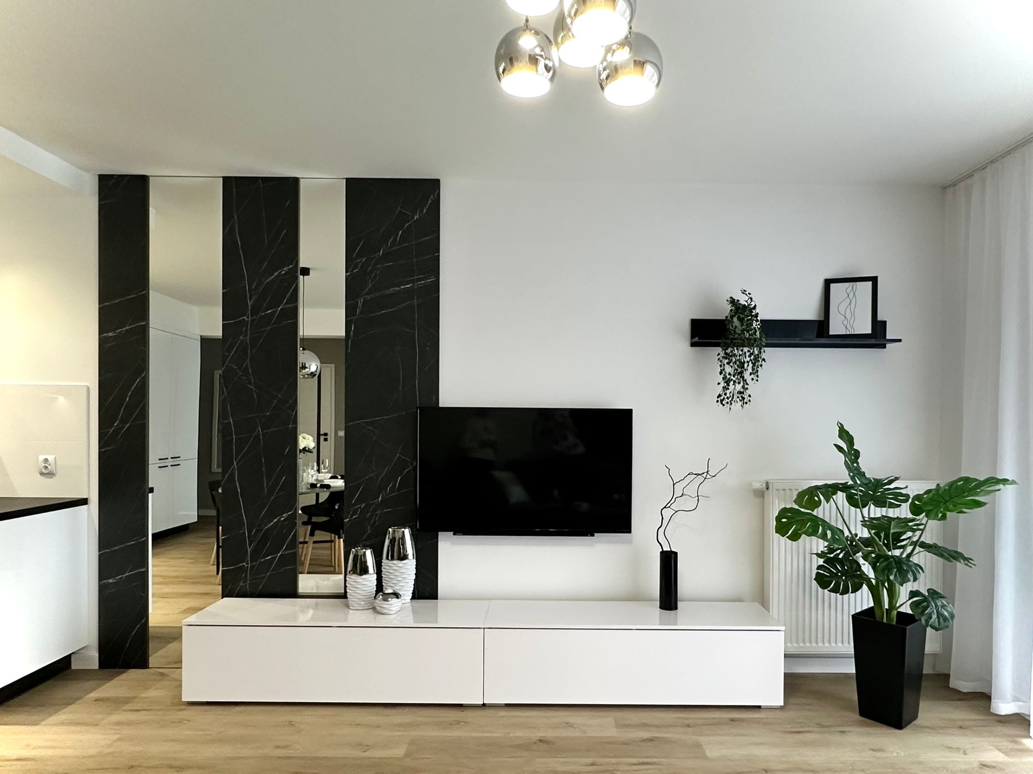 Nowoczesny salon z czarnymi panelami imitującymi marmur, dzielącymi przestrzeń, białą szafką RTV, telewizorem i roślinami doniczkowymi, oświetlony lampą sufitową z chromowanymi kloszami.
