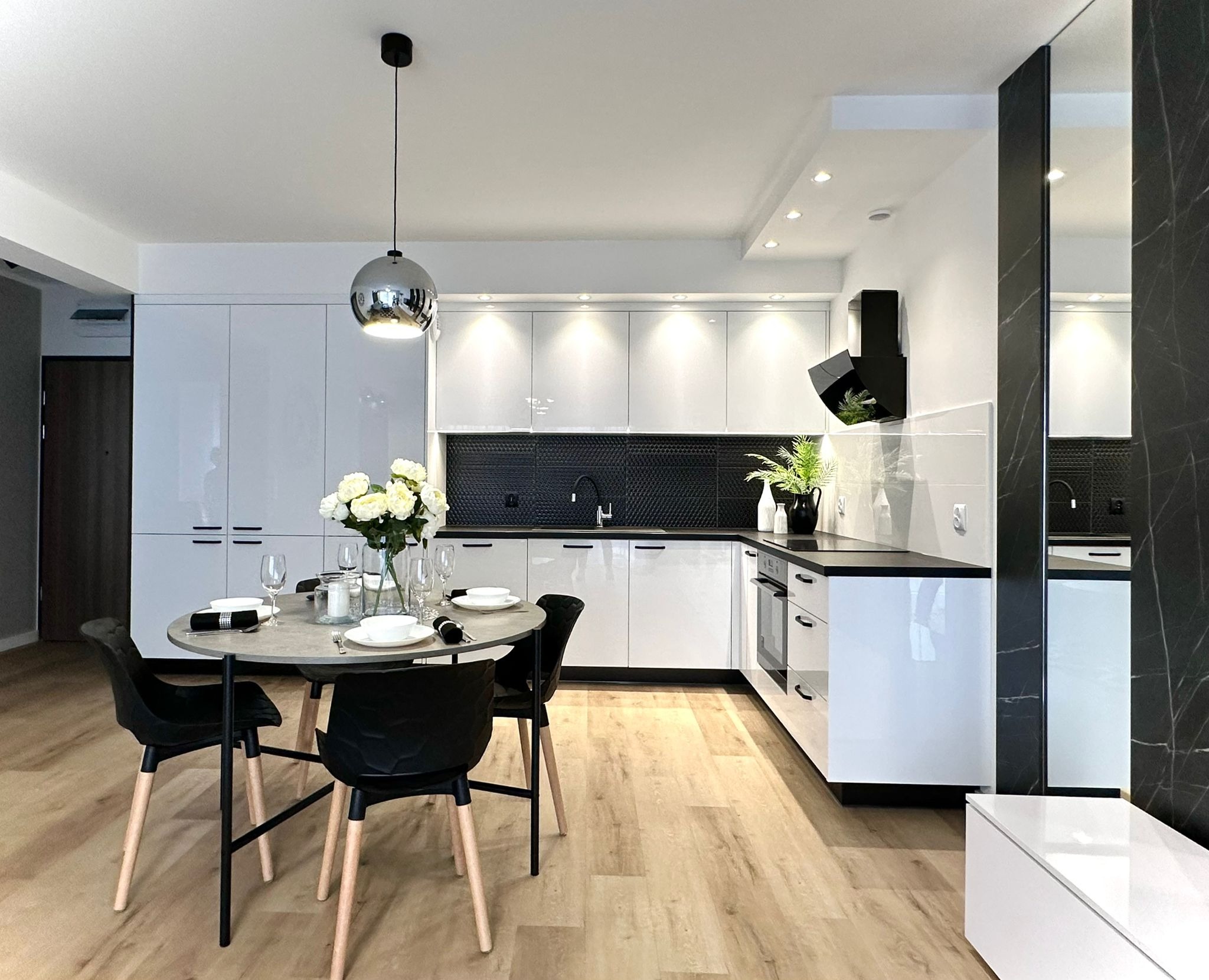 Nowoczesna, jasna kuchnia z białymi, błyszczącymi szafkami, czarnym blatem i heksagonalną mozaiką nad zlewem; okrągły stół jadalny z nakryciem i bukietem białych róż w wazonie, oświetlona...