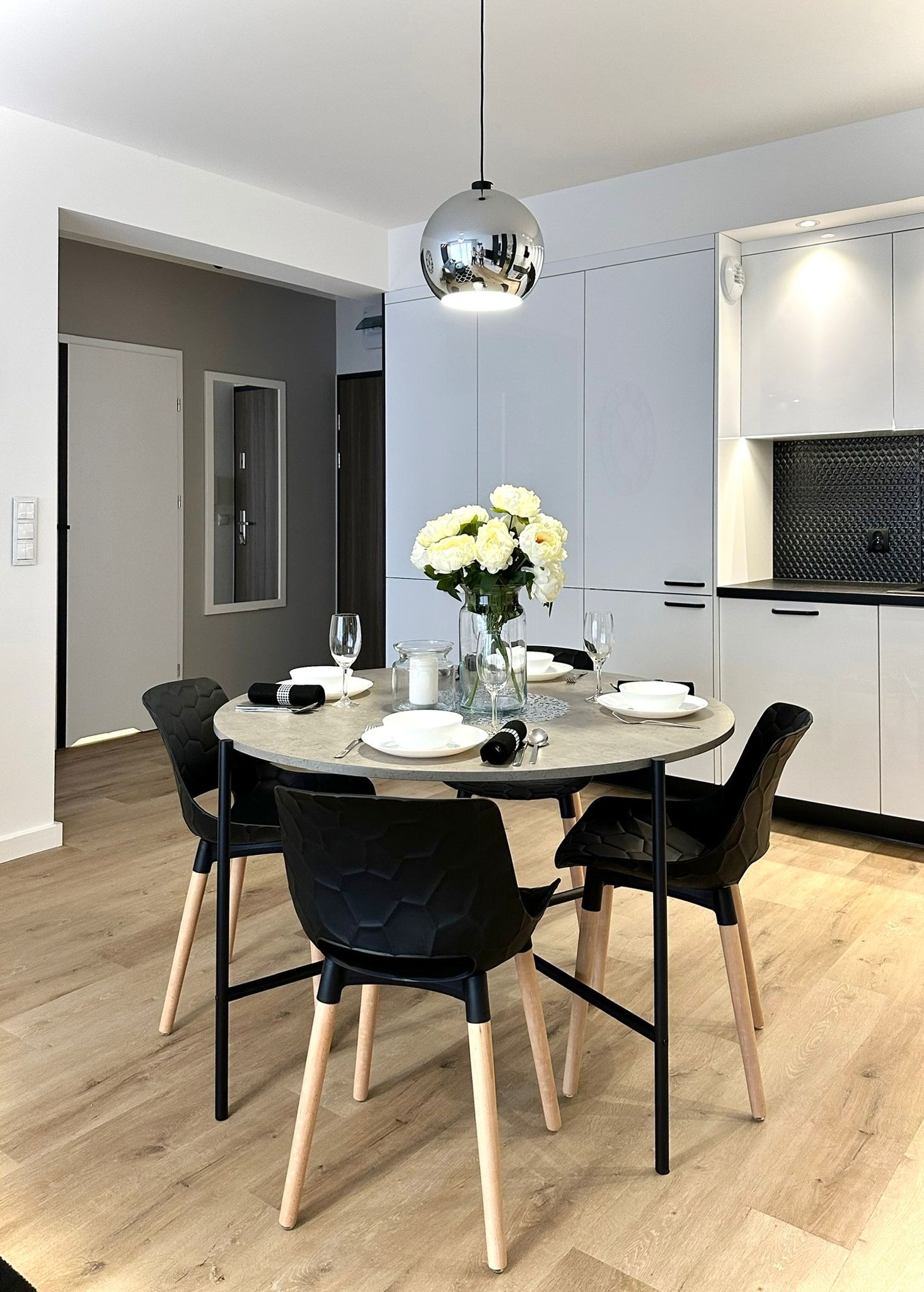 Nowoczesna jadalnia z okrągłym stołem i czarnymi krzesłami na drewnianej podłodze, srebrna lampa wisząca, białe szafki kuchenne w tle, bukiet białych róż w wazonie.
