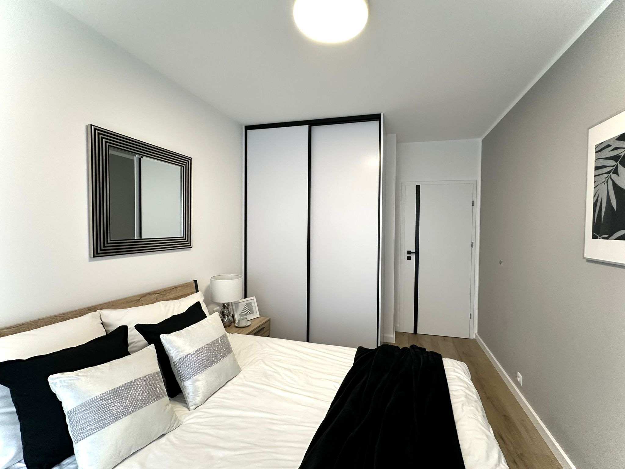 Nowoczesna sypialnia w neutralnych barwach: białe ściany, szara ściana akcentująca, podłoga z paneli, łóżko z czarno-białymi poduszkami i czarnym pledem, szafa przesuwna z czarnymi obramowaniami...